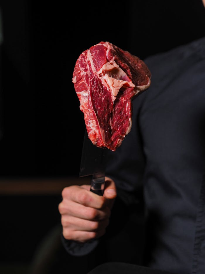why-choose-us A person holds a raw steak on a knife, ready for cooking. Dark indoor setting.